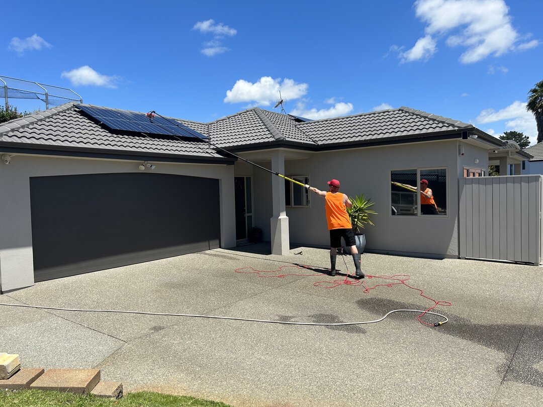 Cleaning a black solar panel on a brown roof with an extended brush.